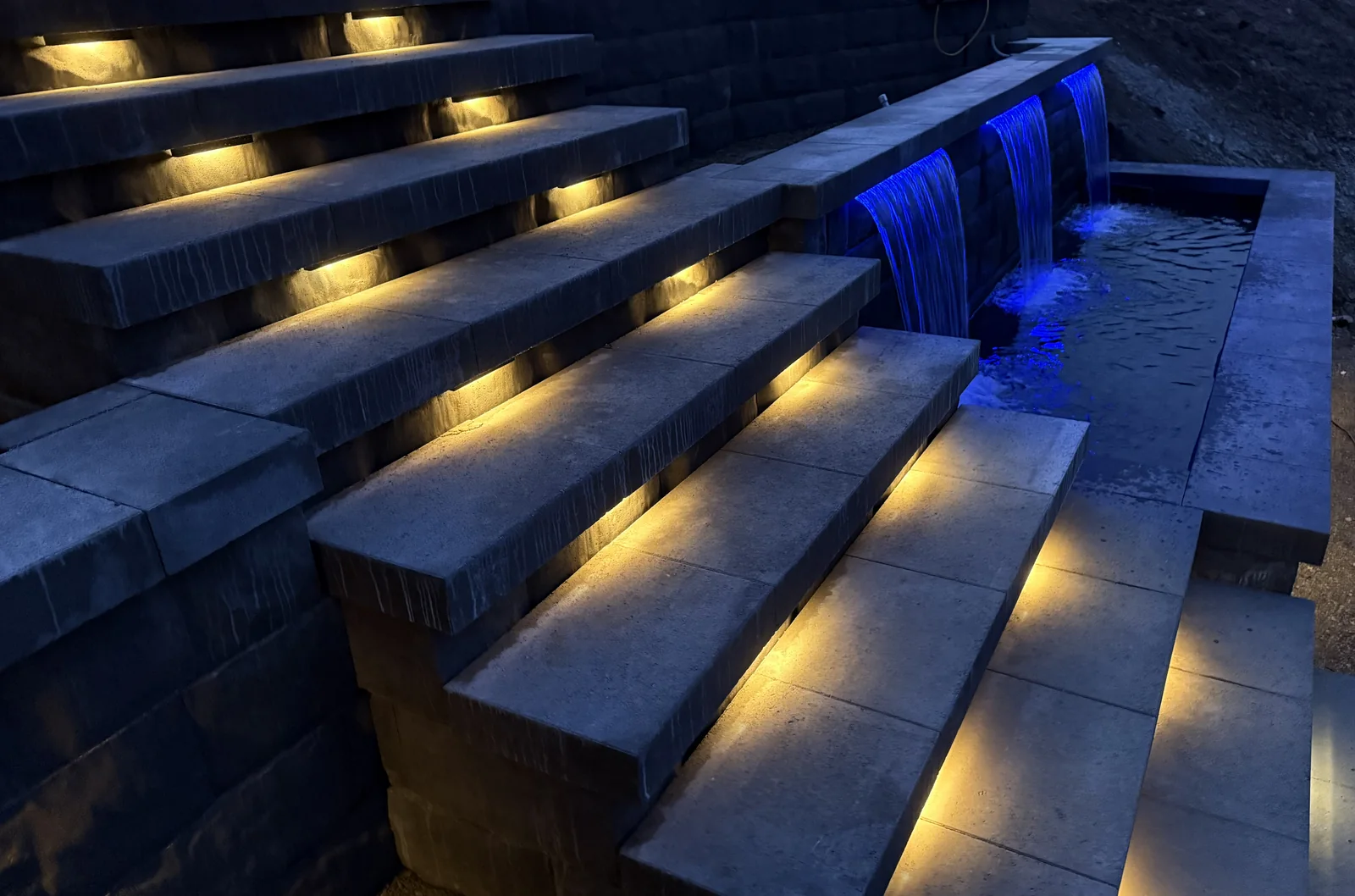 Warm LED step lights running down beside the blue lit water feature