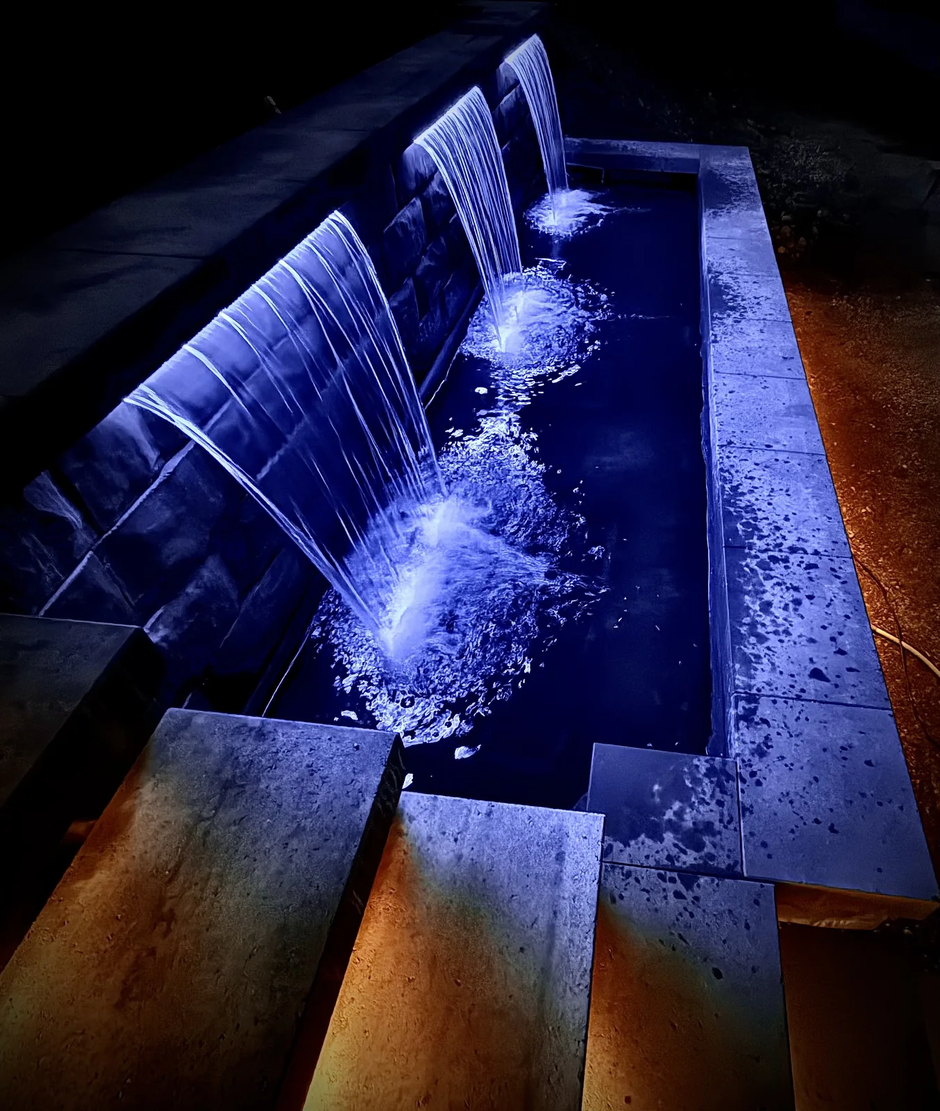 Lit water feature pouring into a basin beside warm lit stone stairs
