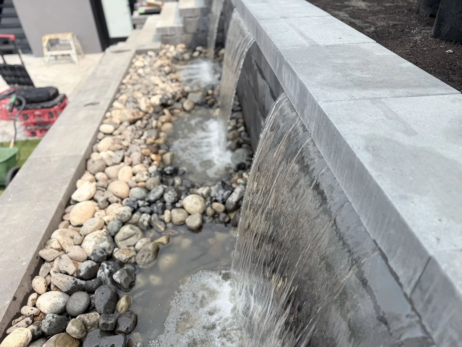 Sheer water feature spilling into a river rock channel beside the pool deck
